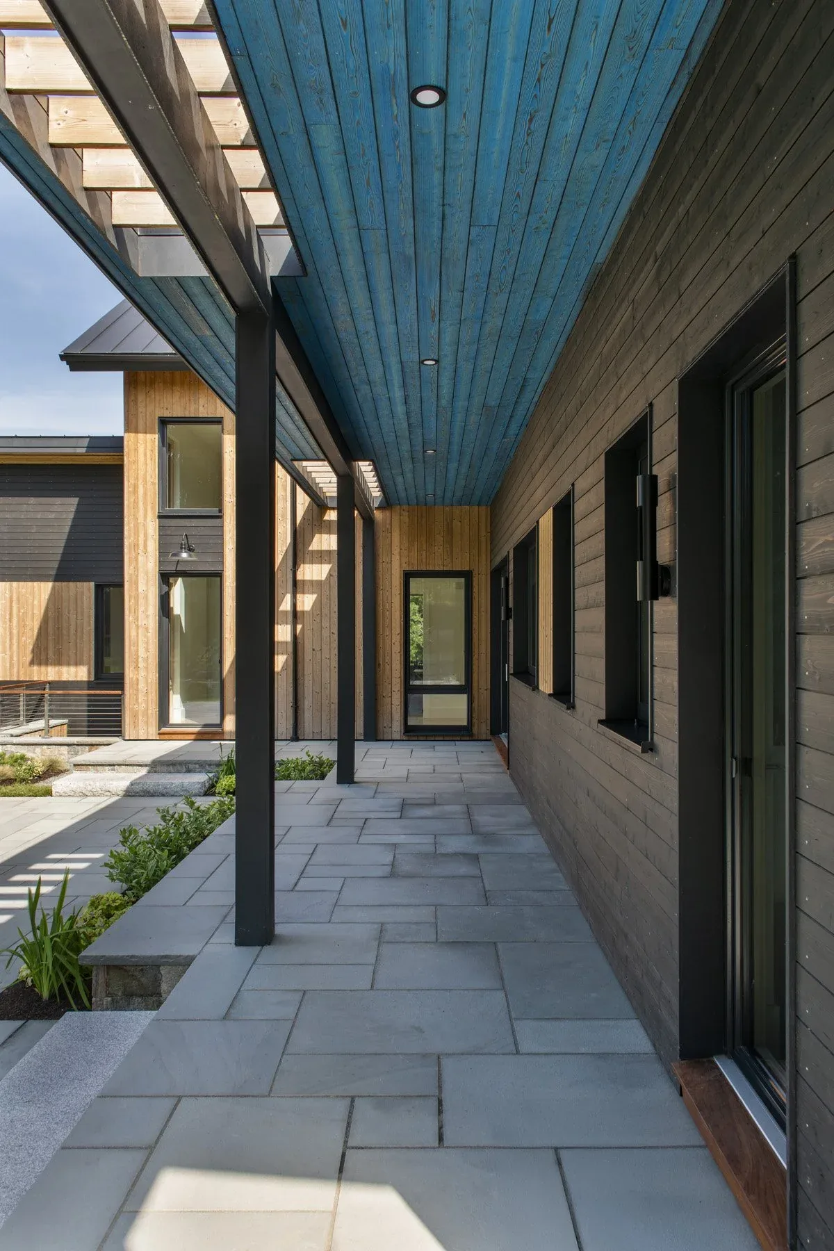 The Stow Passive House features a covered walkway with a blue wood-paneled ceiling and black window frames.