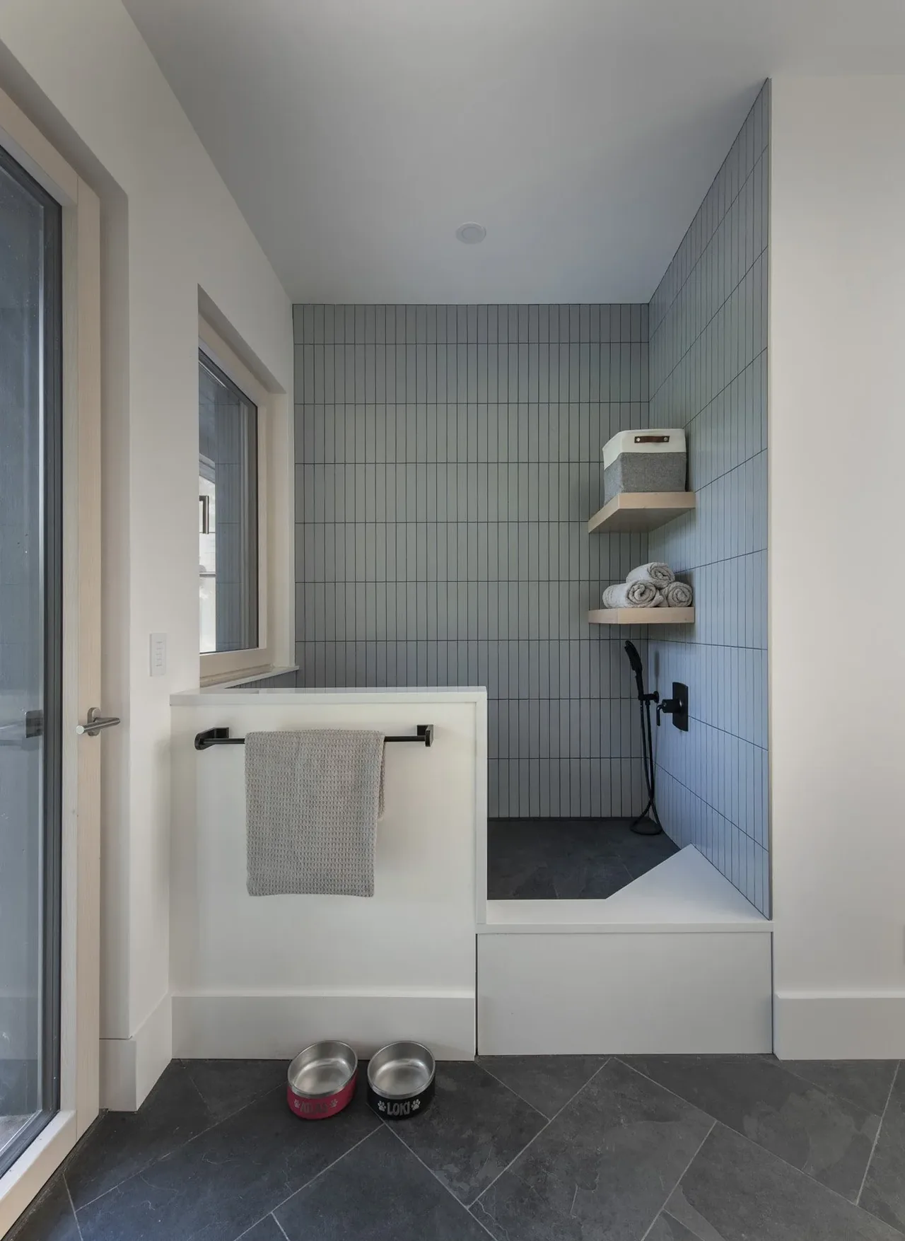 Bathroom shower stall features vertical gray tile, black fixtures, and built-in wood shelves with towels and a storage box.