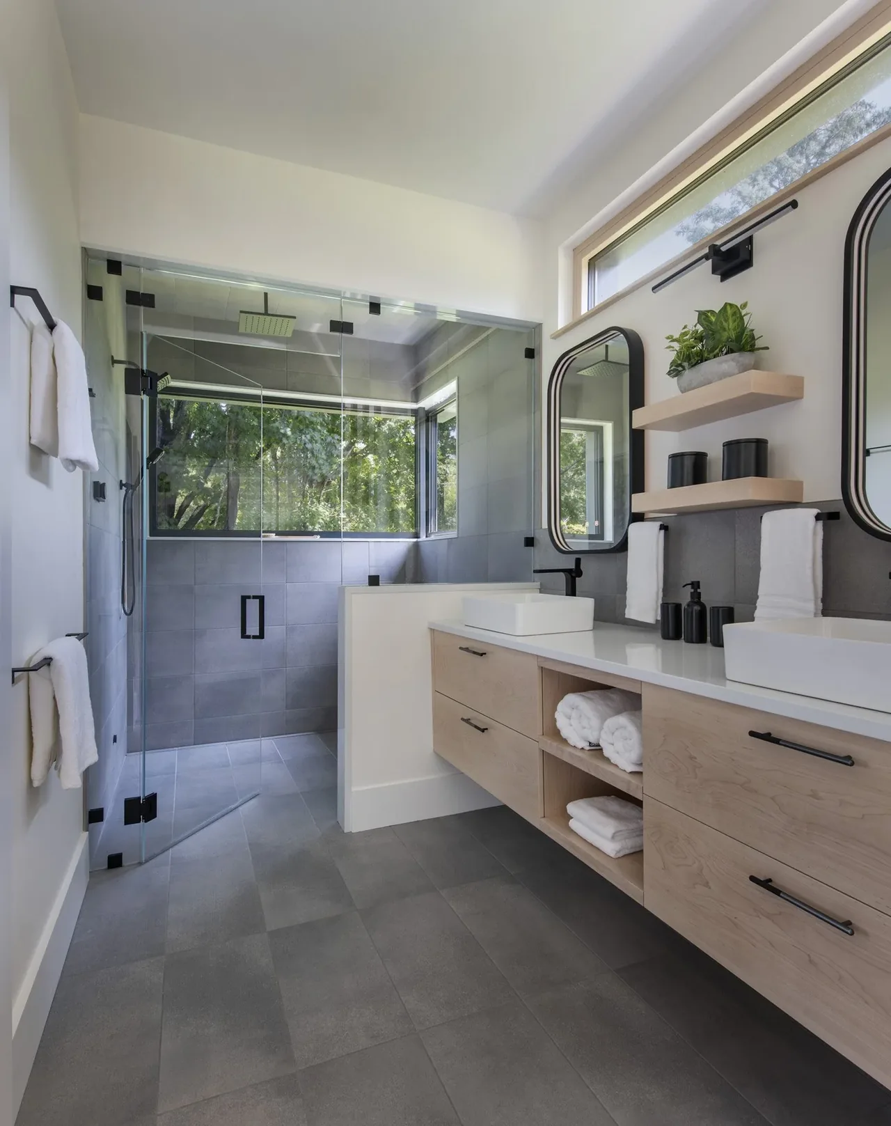 Light wooden vanity with double sinks faces a glass-enclosed shower with a window overlooking greenery.