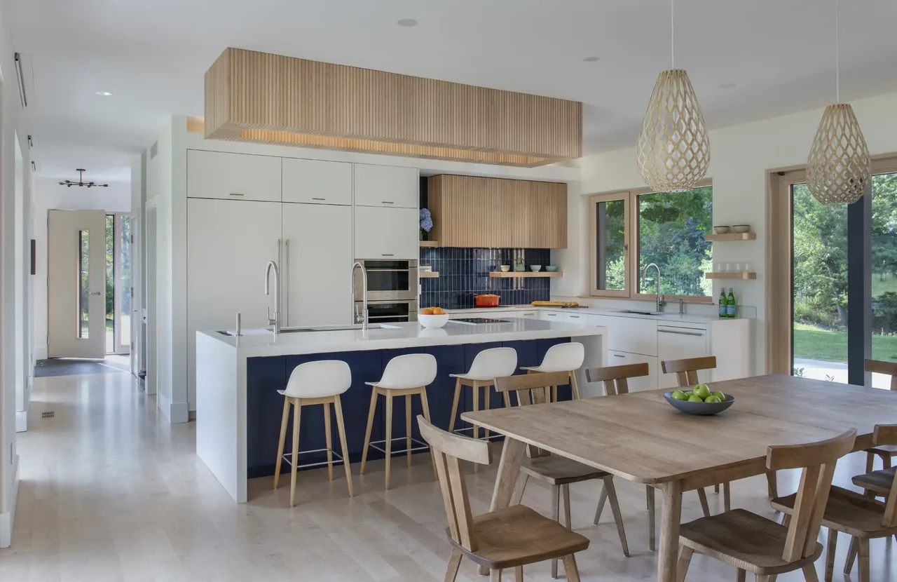 The Stow Passive House kitchen features a white island with bar stools, wood cabinetry, and expansive glazing to the outside.