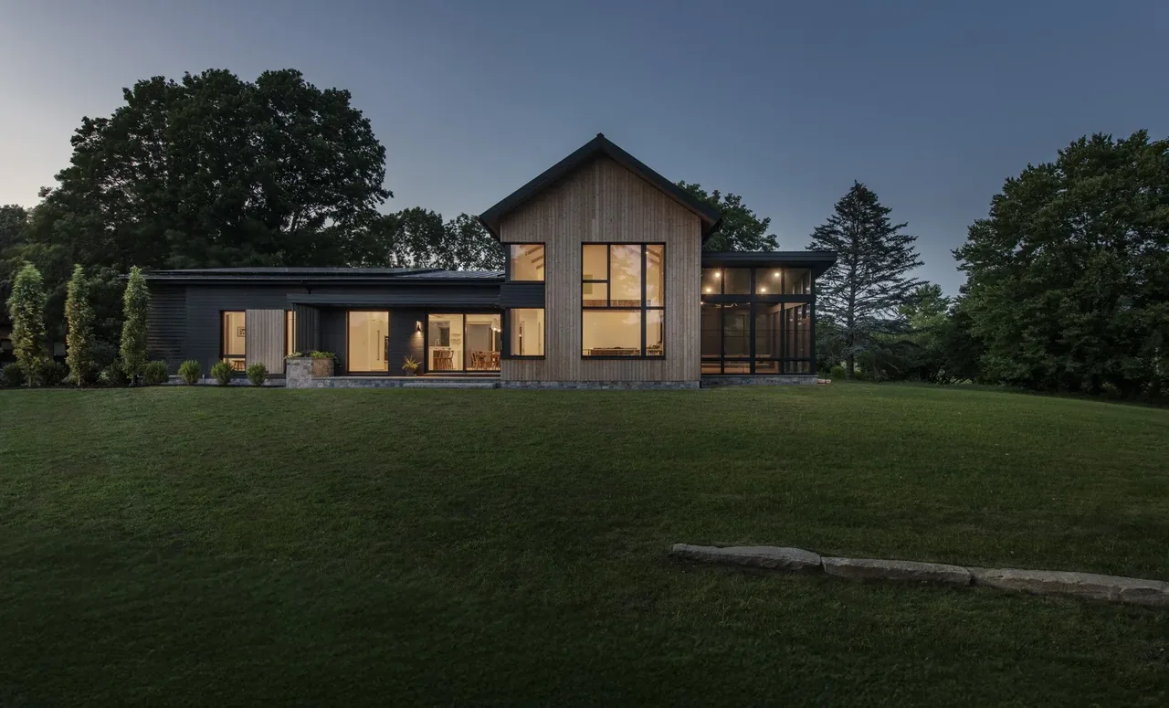 Stow Passive House rear exterior shows wood siding with expansive windows and a screened porch at dusk.