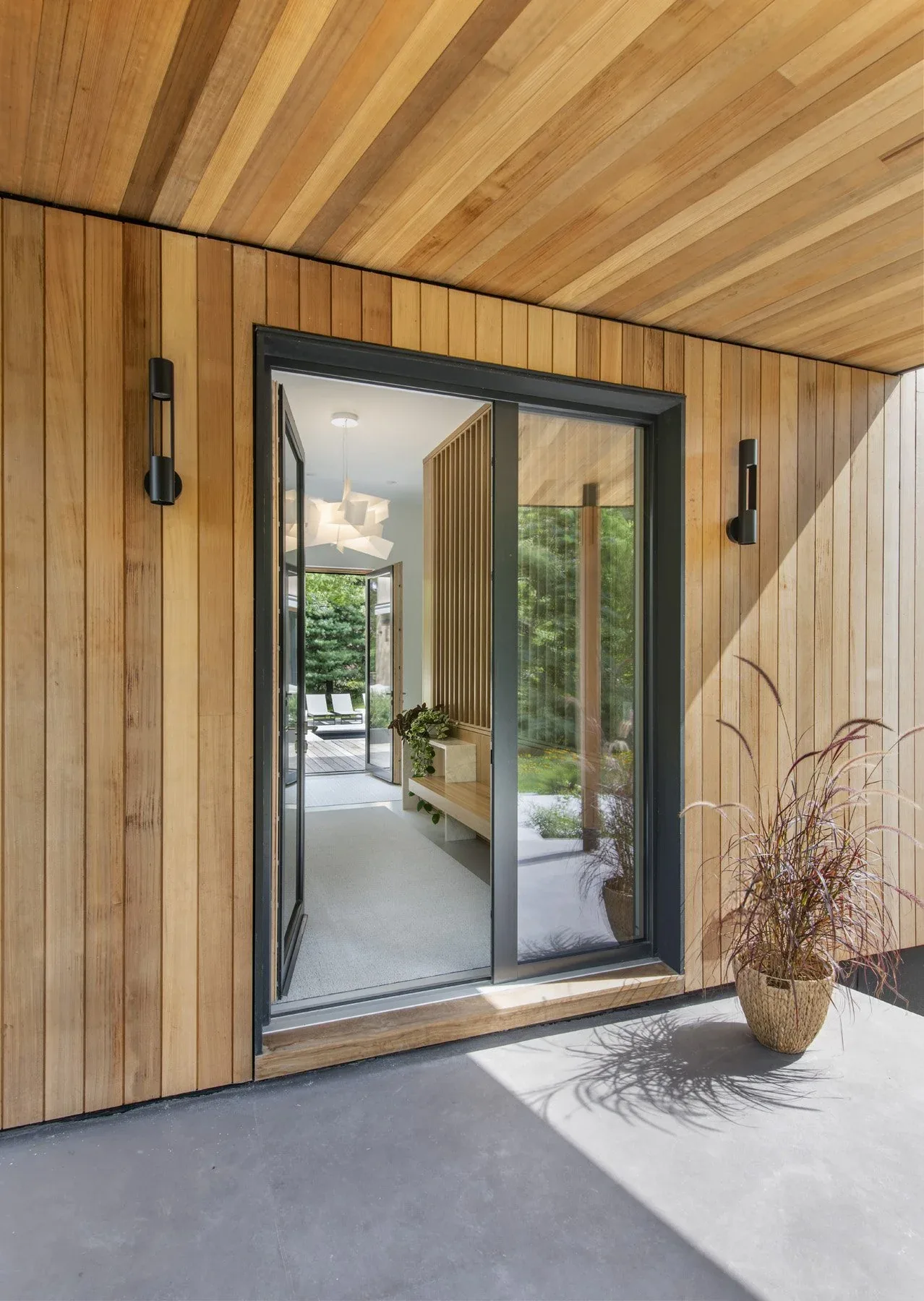 Wood-paneled entry features a black-framed glass pivot door and modern wall sconces, leading into a brightly lit interior.