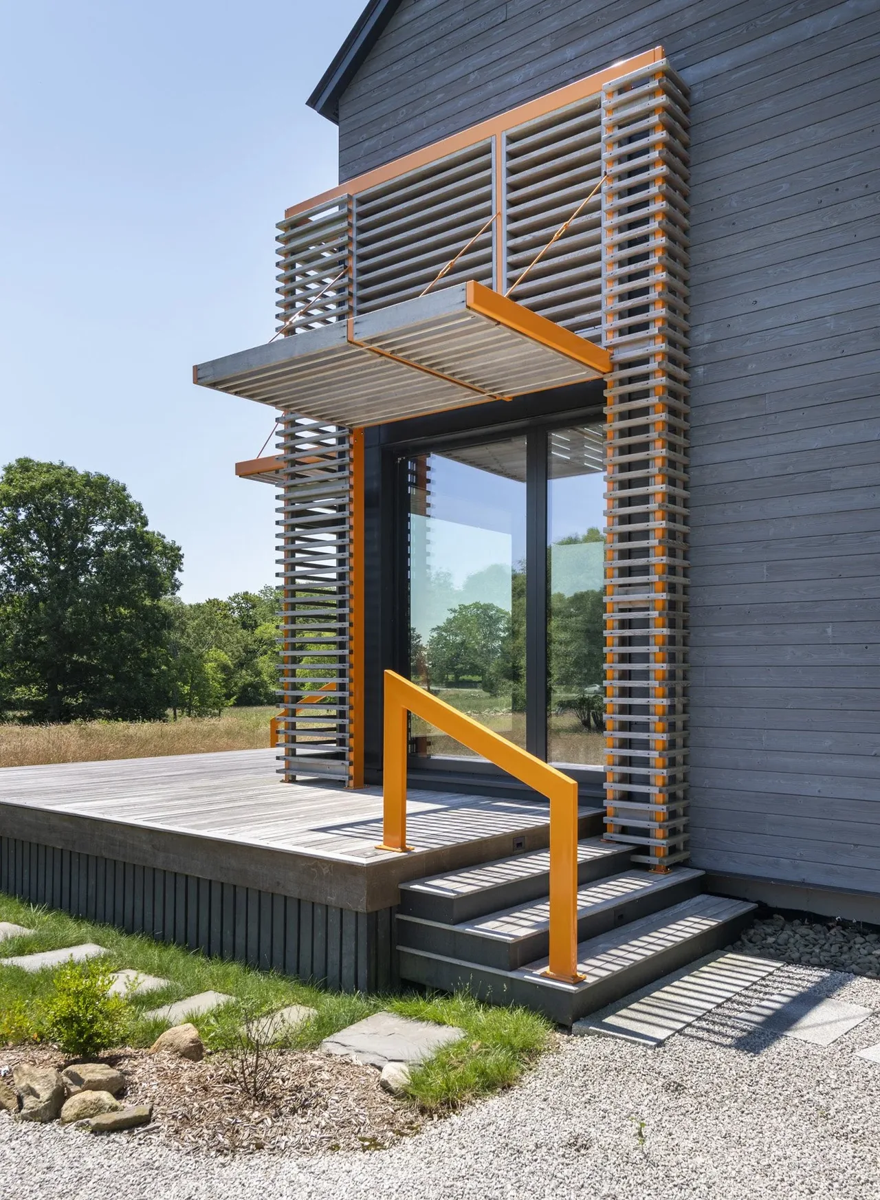 Modern home's exterior entry features a glass door with wood siding and an orange metal sunshade and railing.