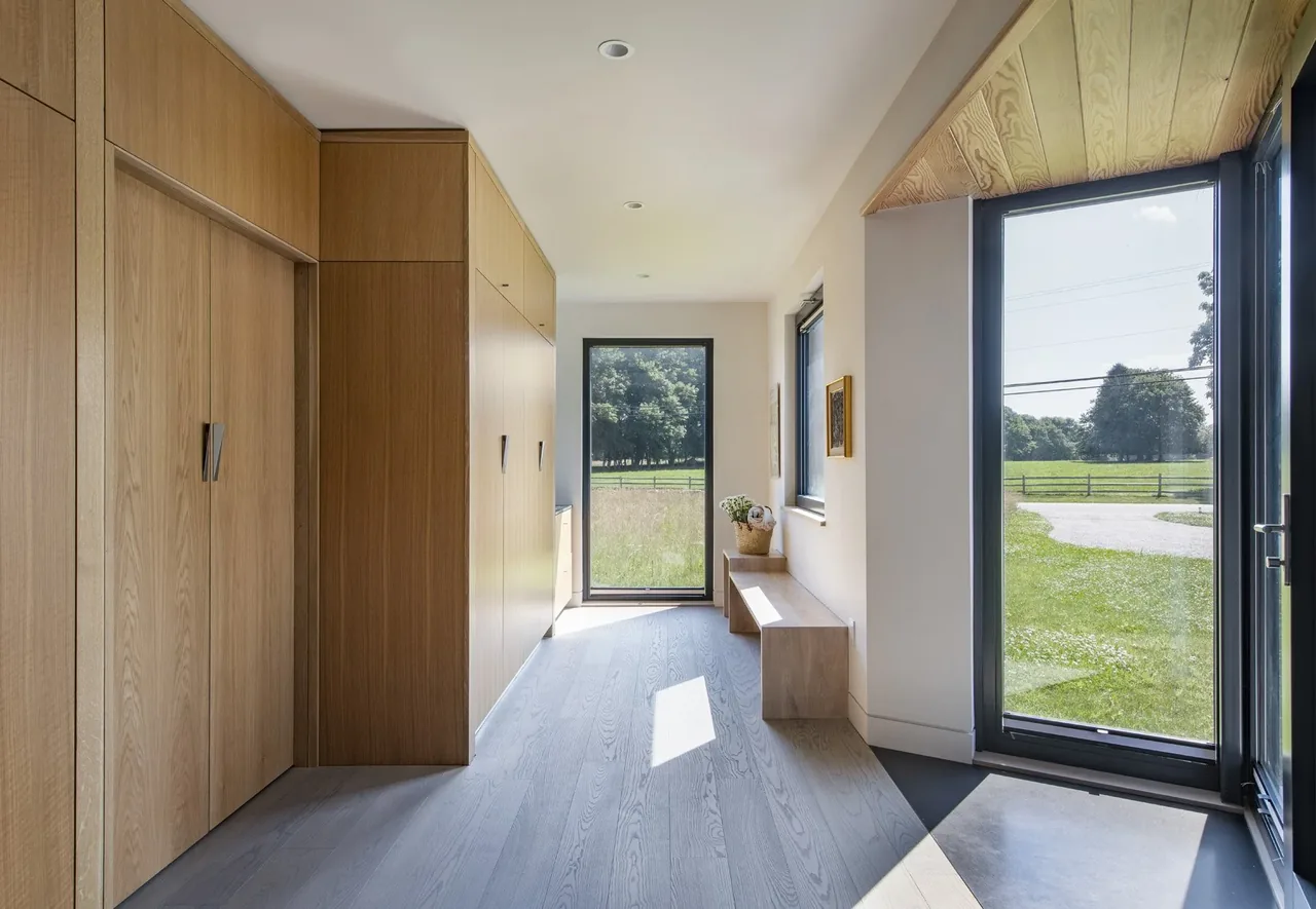 Hallway in the Westport Modern Home features built-in wood cabinetry, a wood bench, and floor-to-ceiling glazing with black window frames.