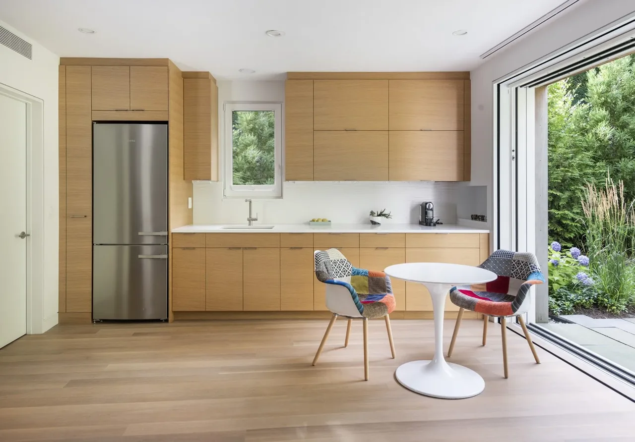 This kitchen features light wood cabinetry, a small dining table with colorful chairs, and a sliding glass door opening to a garden.