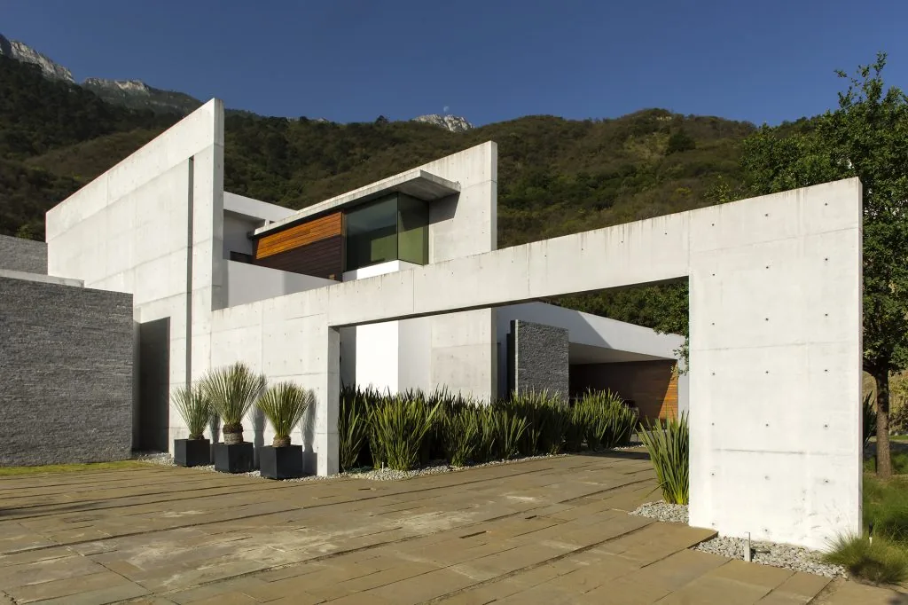 Exterior front view of a modern concrete house with large rectangular architectural elements and landscaping near mountains.