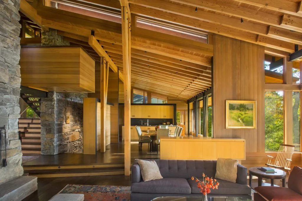 Living and dining area with exposed wood beams and stone wall, showcasing large windows with views of surrounding trees.
