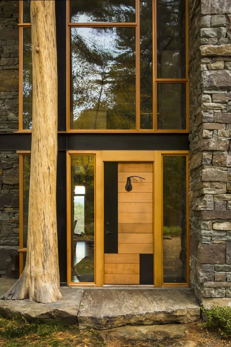 The house entrance features a wooden door flanked by large glass windows, stone cladding, and a vertical log post.