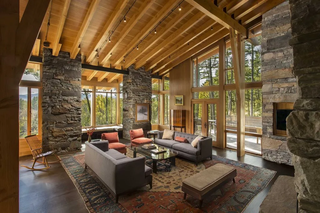 Living area featuring exposed wood ceiling beams, stone pillars, large windows, and modern furniture around a central rug.