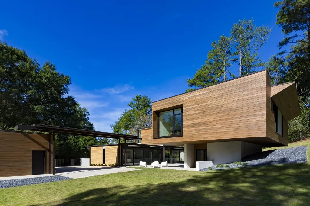 Modern house exterior with wood siding, cantilevered upper volume, and connected carport over concrete driveway.
