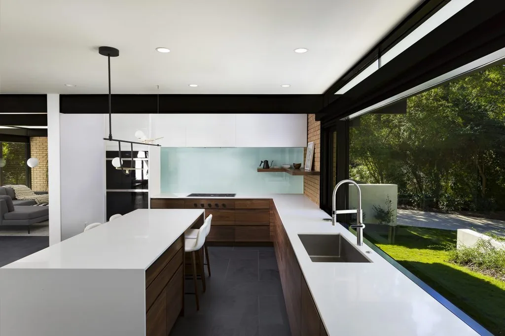 Kitchen features wood cabinetry, white countertops, a glass backsplash, and large picture windows overlooking the yard.