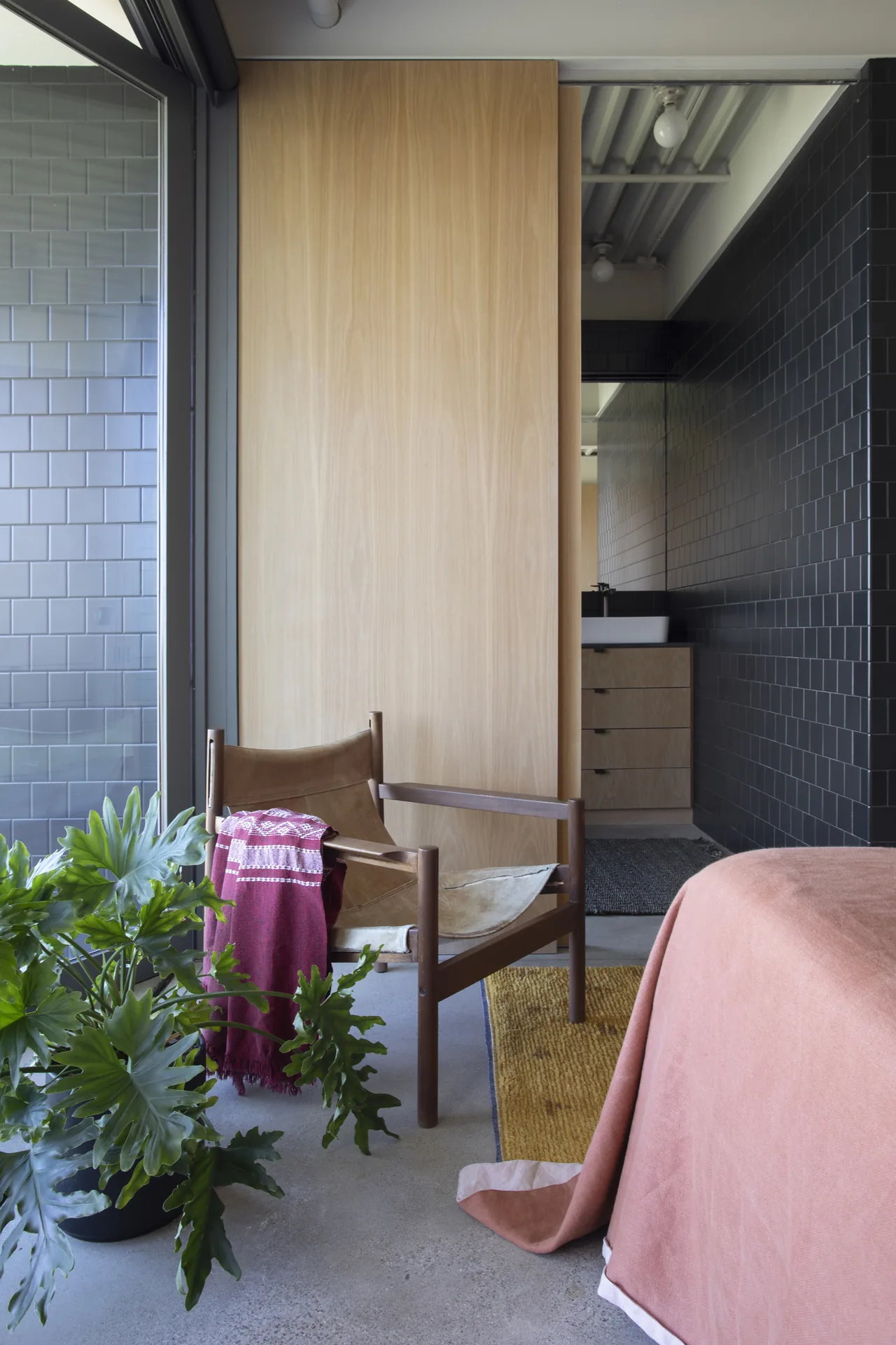 Bedroom with a leather sling chair, coral bedding, and visible bathroom with black tile walls at Olancha House.