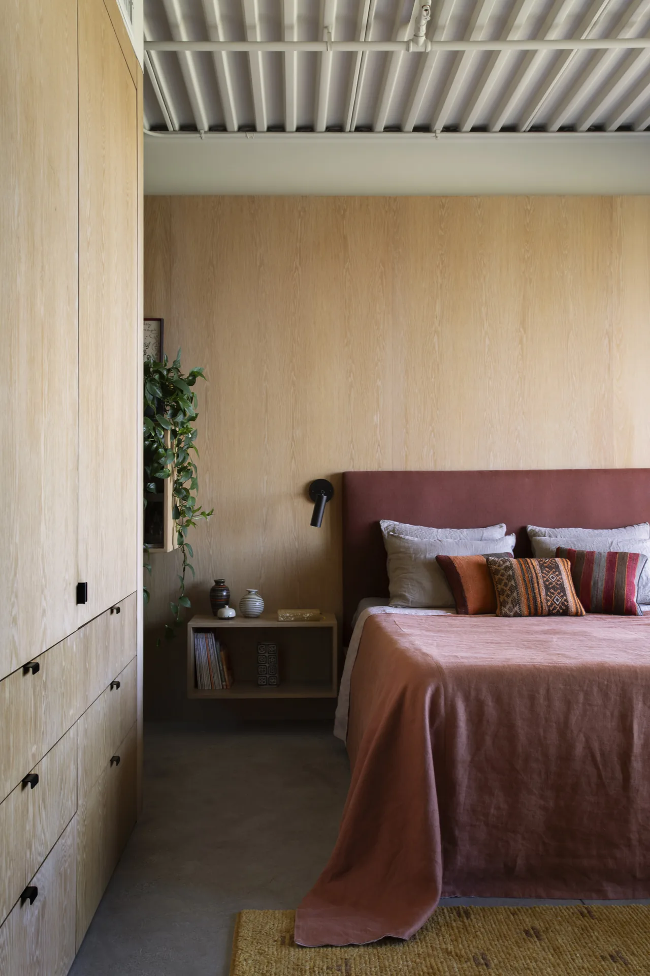 Bedroom with wood-paneled walls, a rust-colored bedspread and headboard, and built-in storage cabinets by Anonymous Architects at Olancha House.