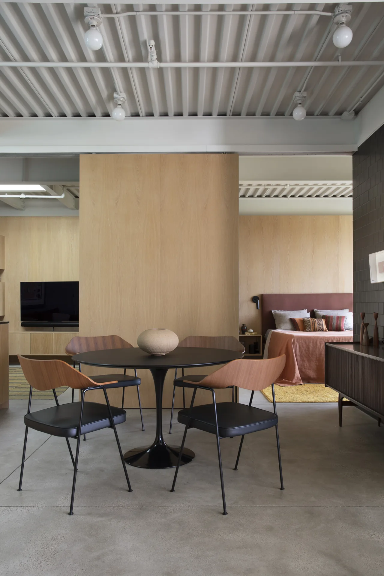The Olancha House dining area features a modern round black table and chairs, with a visible bedroom space showcasing coral textiles and wood walls.