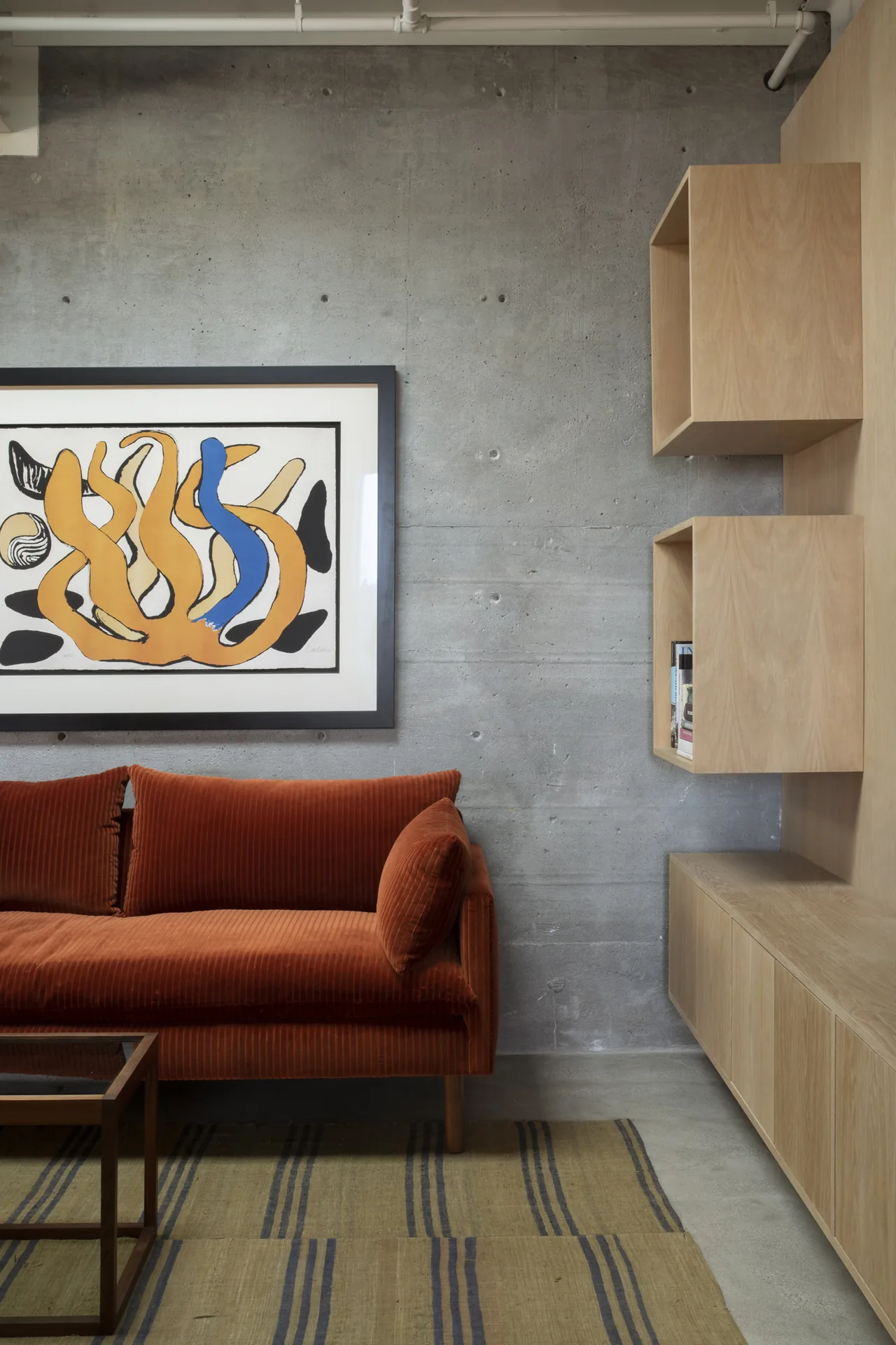 Living room in Olancha House features a rust-colored sofa, concrete wall, and minimalist wood shelving with abstract artwork.