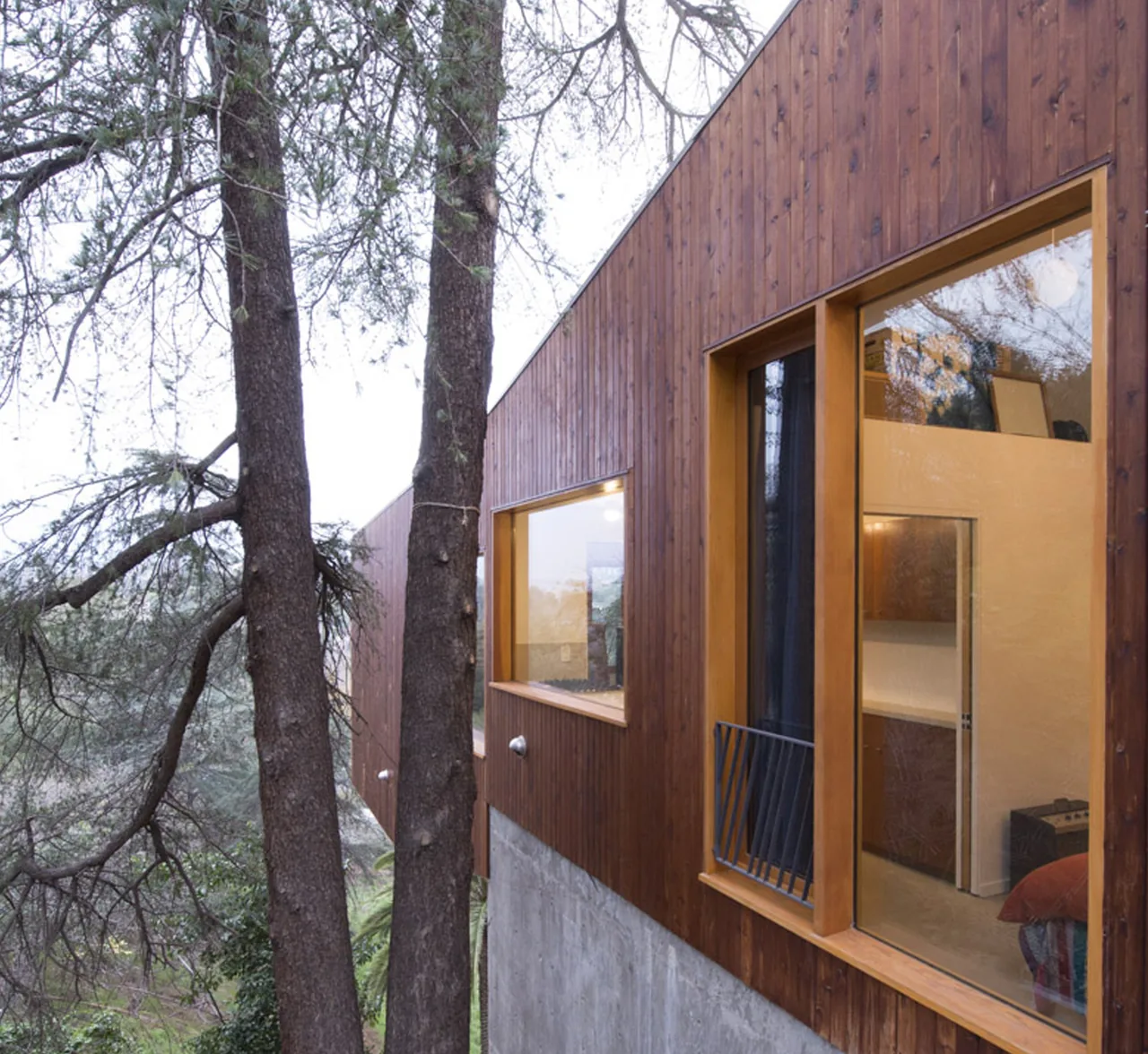 Modern residence with vertical wood siding and large window openings overlooking wooded surroundings.