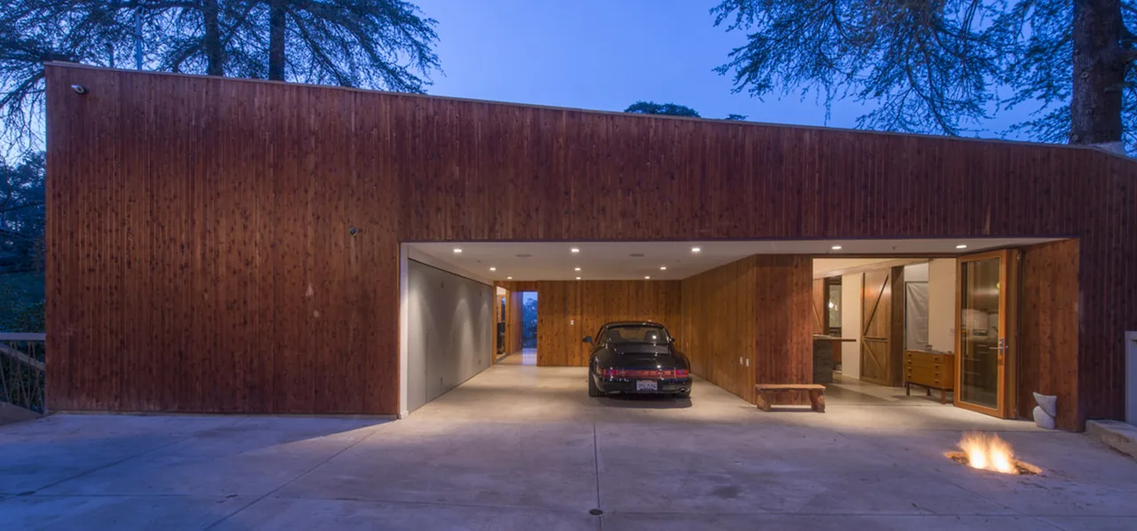Hilltop house garage with wood siding, illuminated at dusk, featuring a parked car and a small fire pit on the driveway.