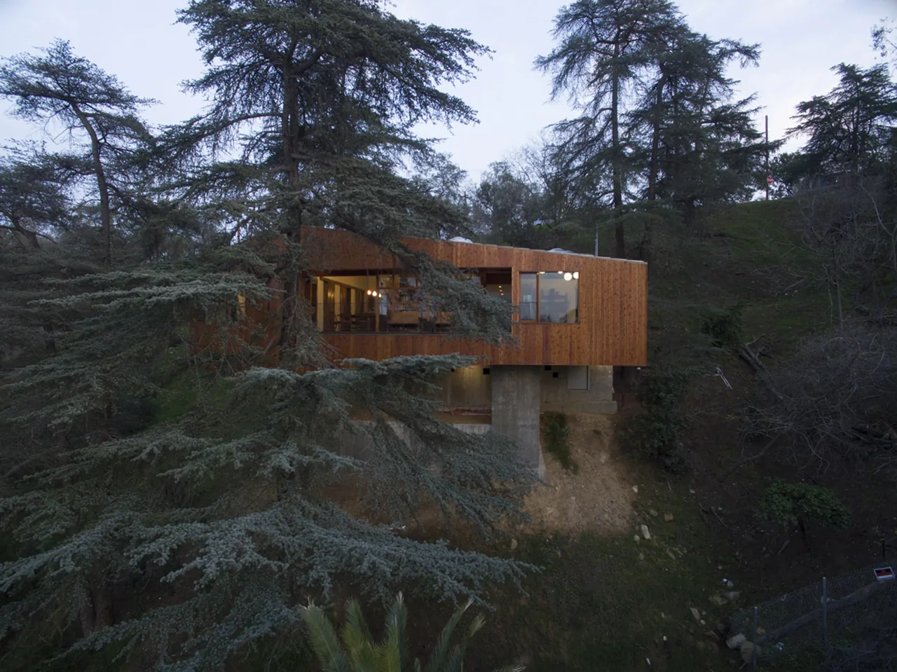 Modern house with wood siding cantilevered on a hillside, featuring large windows and exposed concrete supports.