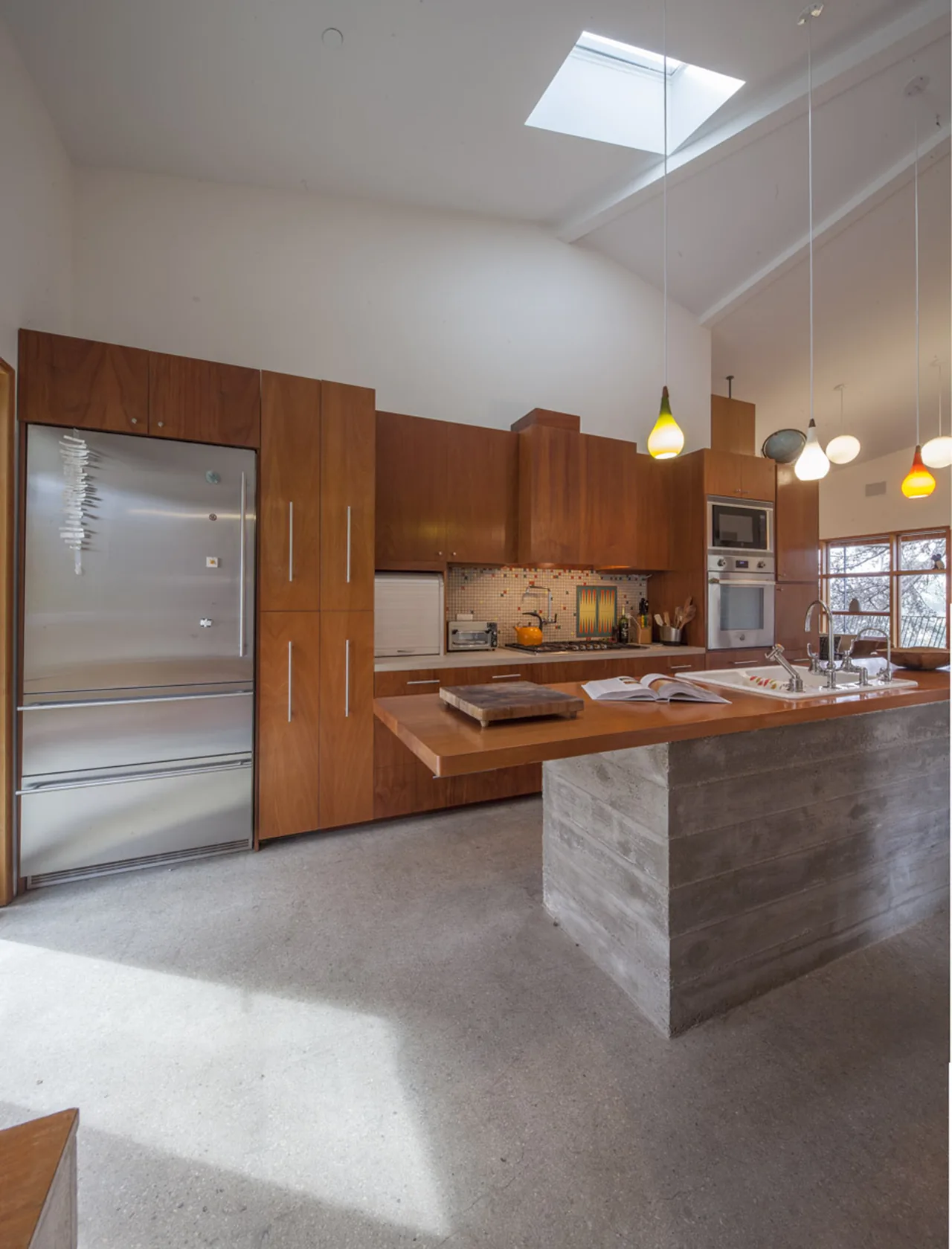 A kitchen featuring warm wood cabinets, a concrete island, skylight, and pendant lighting with colorful shades above the counter.