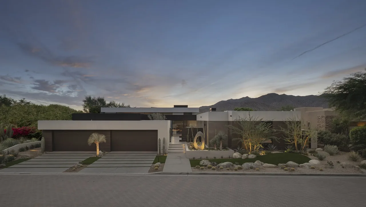 Modern flat-roof house with desert landscaping and three-car garage, set against mountain backdrop at sunset.