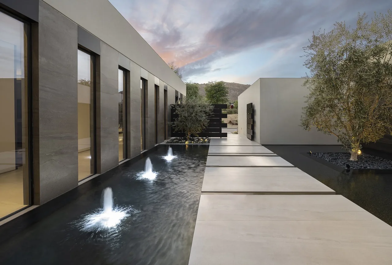 Concrete stepping stones over water fountains create a modern walkway beside a building with vertical windows and mountain backdrop.