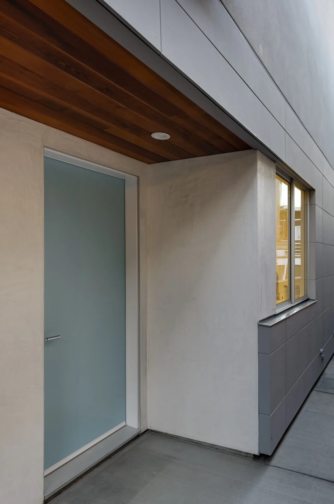 Frosted glass entry door with wood-paneled ceiling in a sleek concrete exterior corridor.