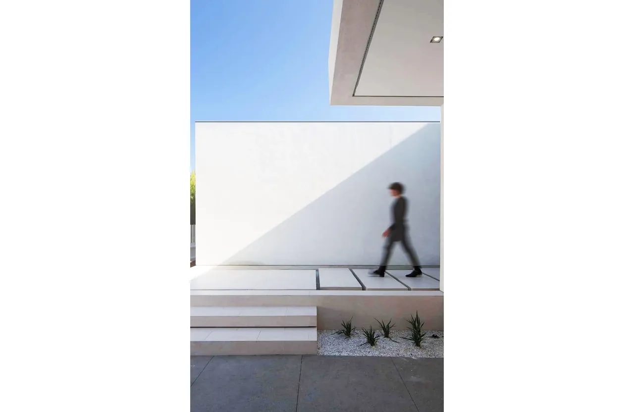 Minimalist white exterior wall with stepped platform and planted gravel bed in sunlight.