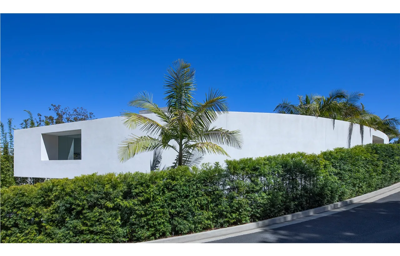 Curved white stucco exterior wall with a rectangular window set back, surrounded by manicured hedges and palm trees.