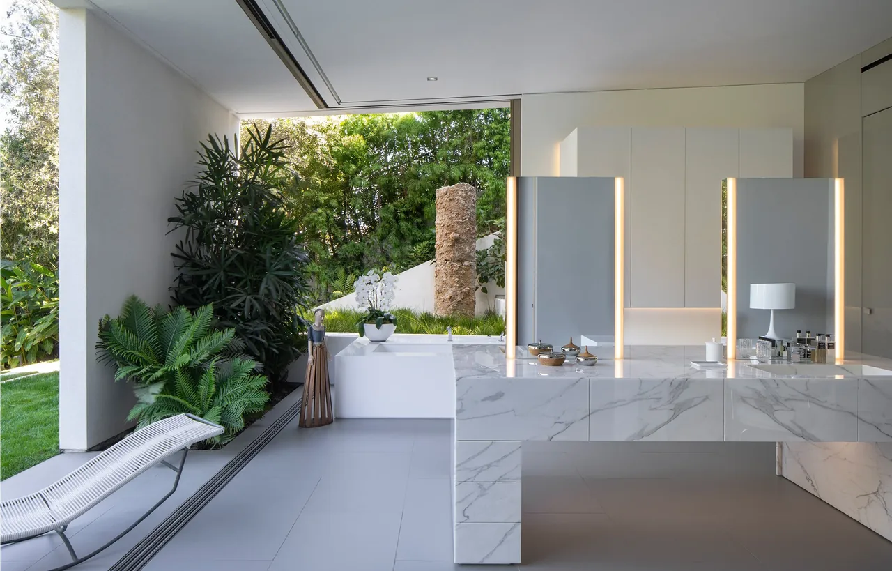 The aMahouse bathroom features a marble vanity with illuminated mirrors and open access to a landscaped green area.