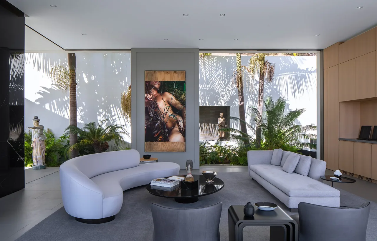The living room of aMahouse features a curved white sofa, a gray sofa, and large windows showcasing lush landscaping outside.