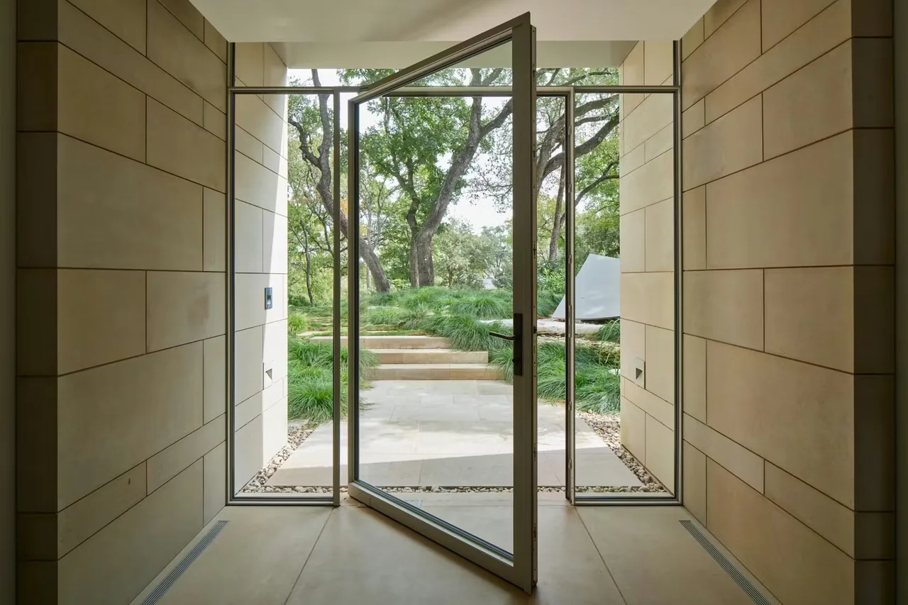 Pivot door with black window frames opens from a stone-floored entry to an outdoor space with stone steps and greenery.