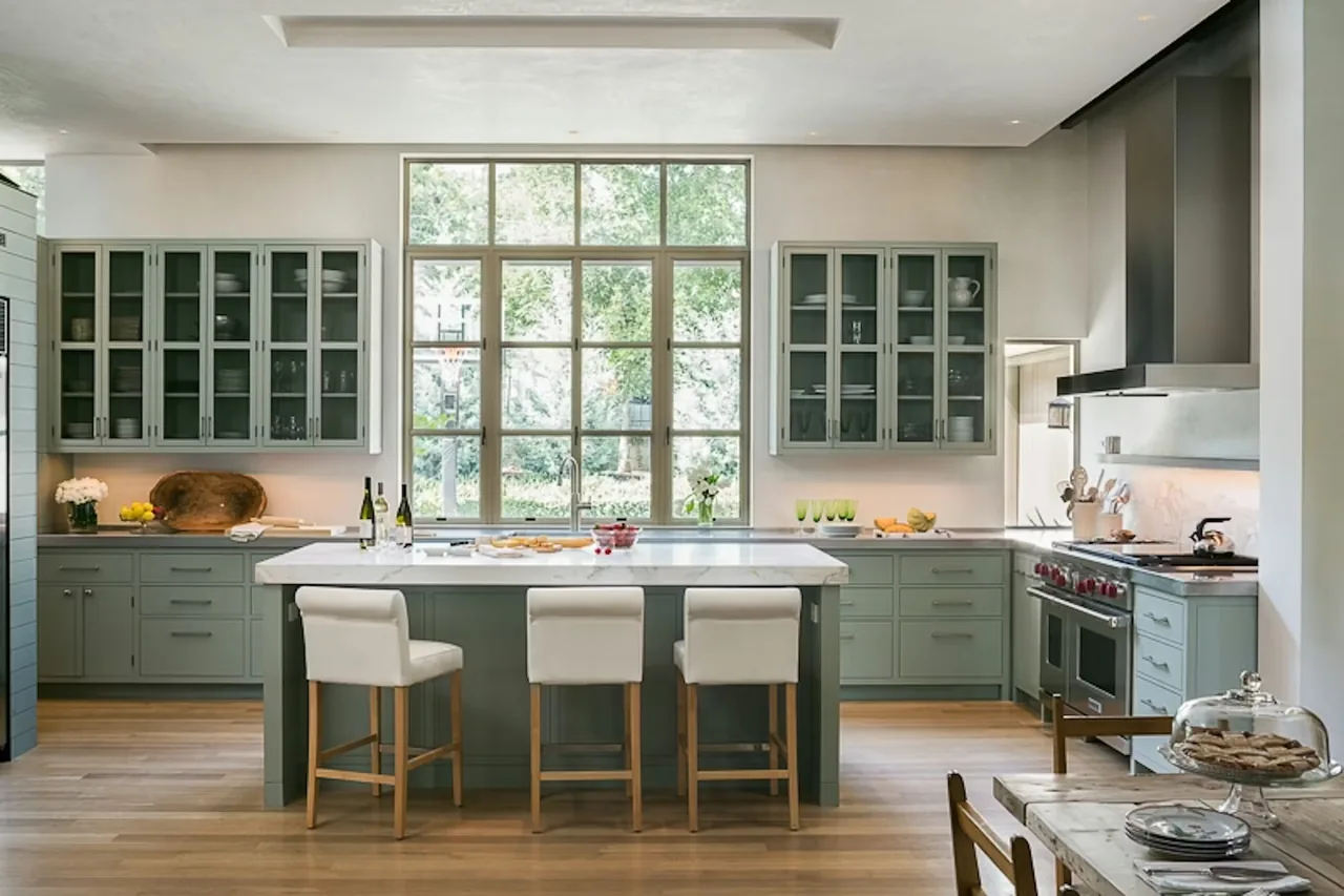 Tarryhill Residence kitchen features sage green cabinetry, large grid window, and white marble island with three stools.