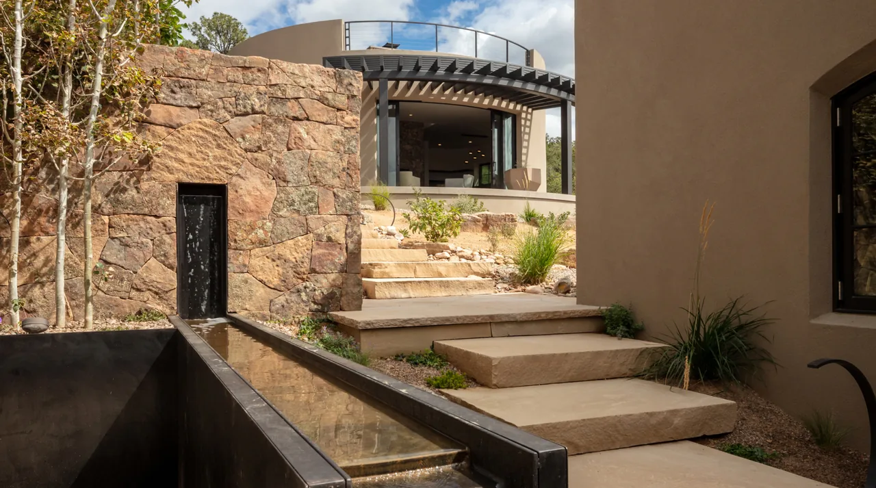 Modern exterior showing curved stucco walls, arched metal awning, stone retaining wall, and terraced landscaping.