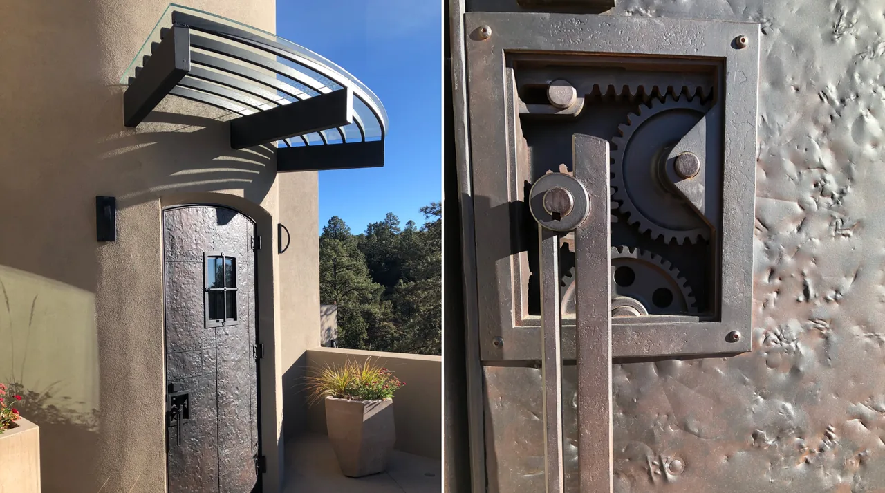 Arched hammered metal exterior door with small glazed window and decorative exposed gears, shaded by curved metal awning.