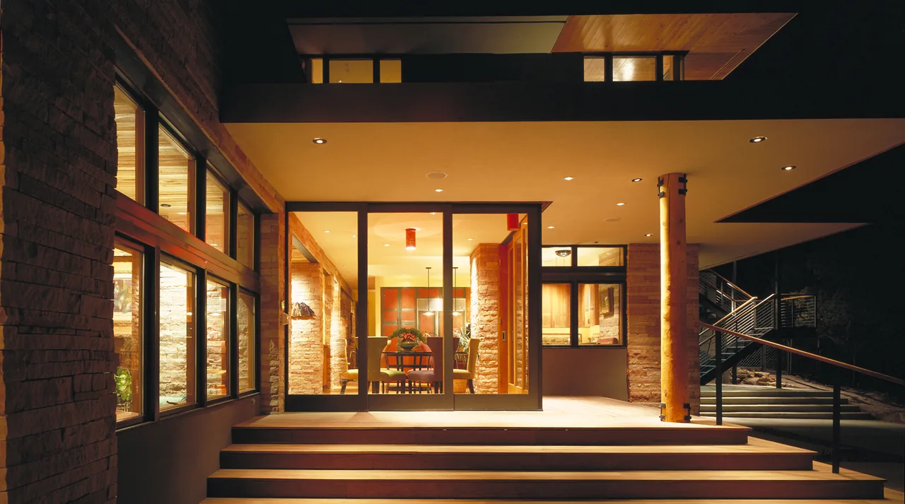 Exterior night view of a modern multi-level home with stone walls, large glass windows, and metal railings on the steps.