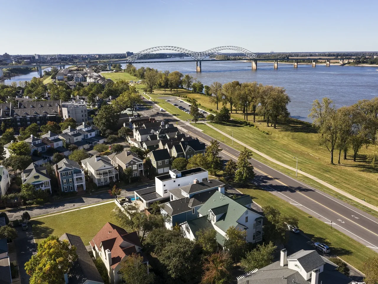 Riverfront neighborhood with mixed residential styles along a street and a large arch bridge across the river in the distance.