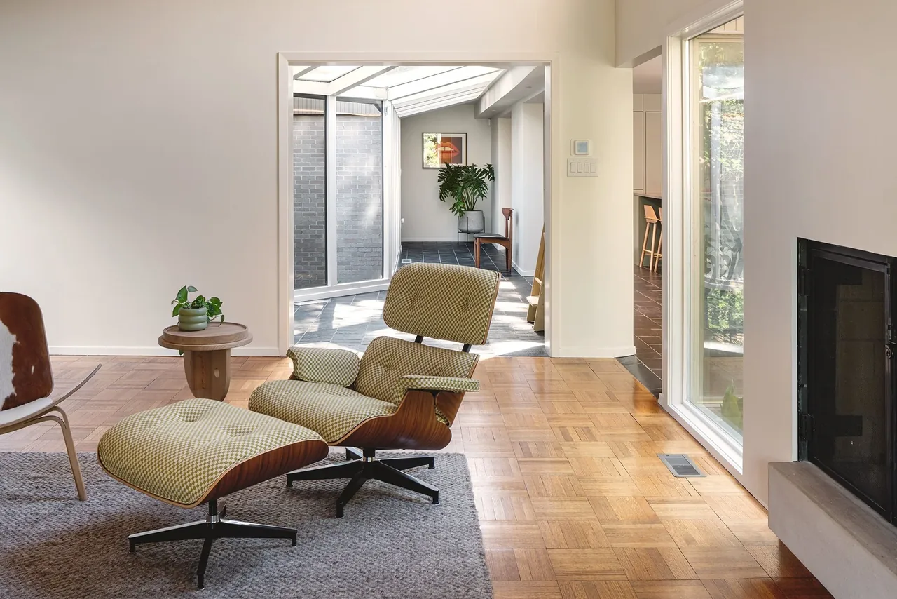 The Russell Residence living space features parquet flooring, a lounge chair with ottoman, and large windows overlooking a patio.