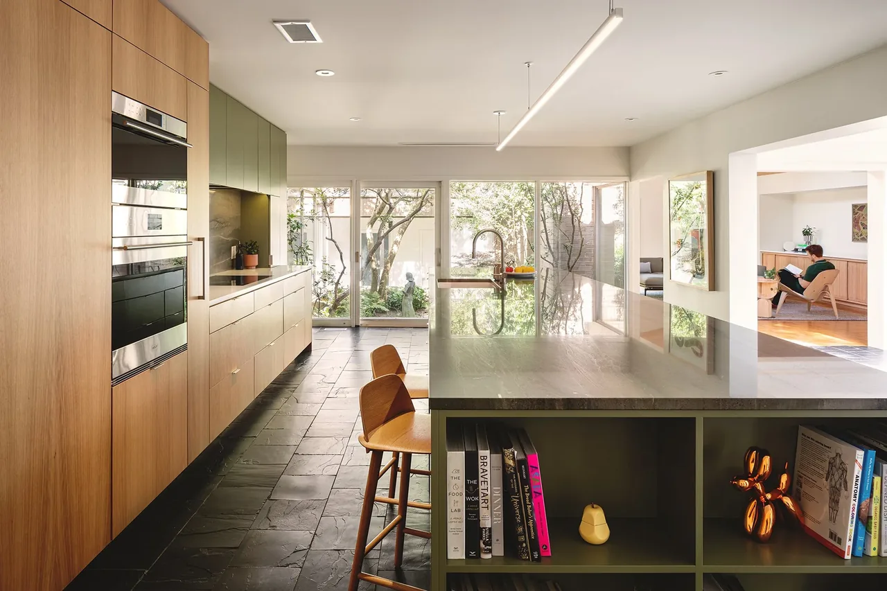 Modern kitchen with wood cabinets, a dark stone floor, and a large island featuring an open shelf and stools, illuminated by a linear light.