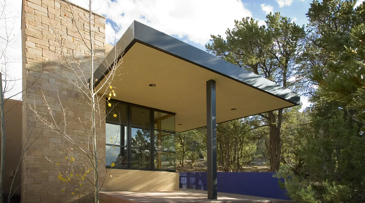 The Lehman project features a flat roof over a covered patio, stone walls, black window frames, and a blue privacy fence near a wooded area.