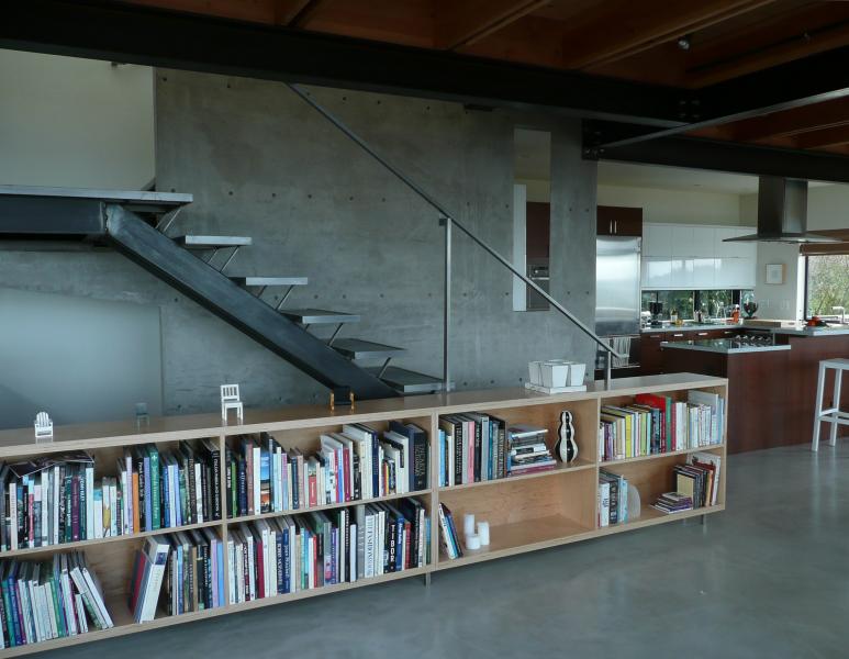 Modern interior featuring a metal open-riser staircase, concrete wall, and built-in bookshelf with an adjacent kitchen space.