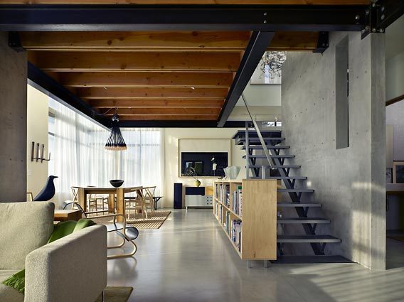 Open floor plan with built-in bookshelf under steel stairs and exposed wood ceiling beams.