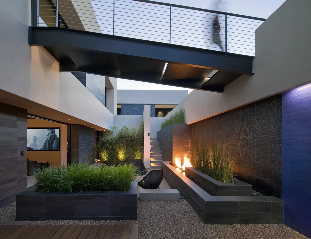 The tresARCA project by Assemblage Studio features an open courtyard with a fire feature and a metal pedestrian bridge overhead.