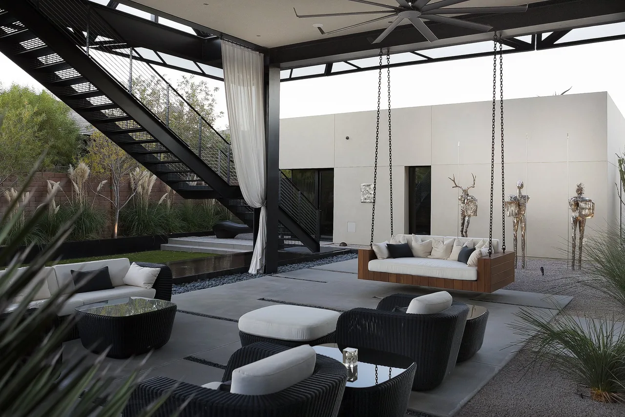 Outdoor living space with suspended wooden sofa and metal stairs opening to a minimalist courtyard featuring metal sculptures.