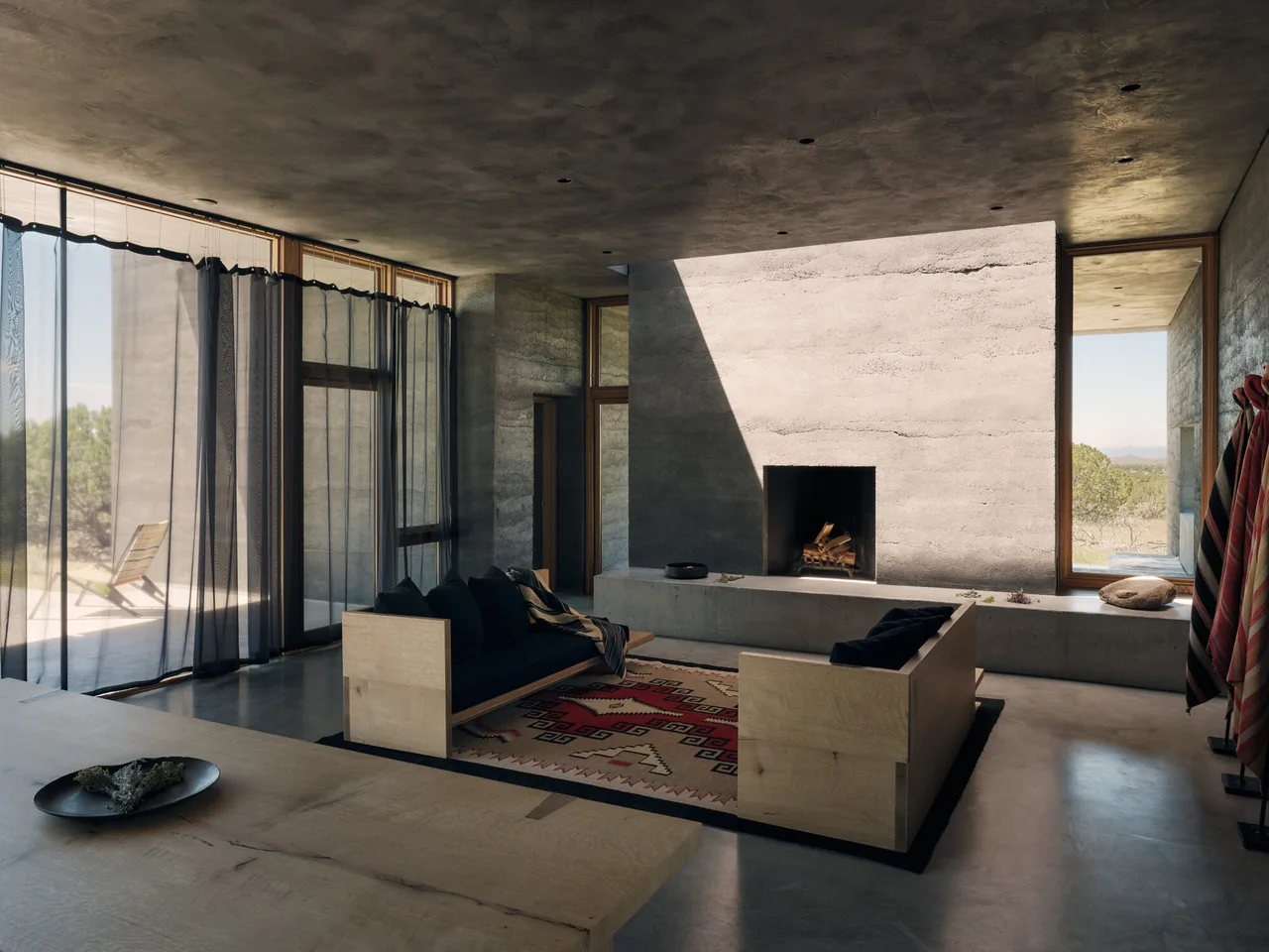 Living room of Sombra de Santa Fe with textured concrete walls and a built-in recessed fireplace by floor-to-ceiling glazing.
