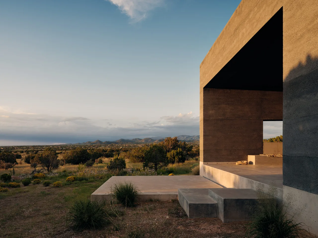 Sombra de Santa Fe features a modern concrete patio with built-in seating facing a desert landscape and distant mountains.