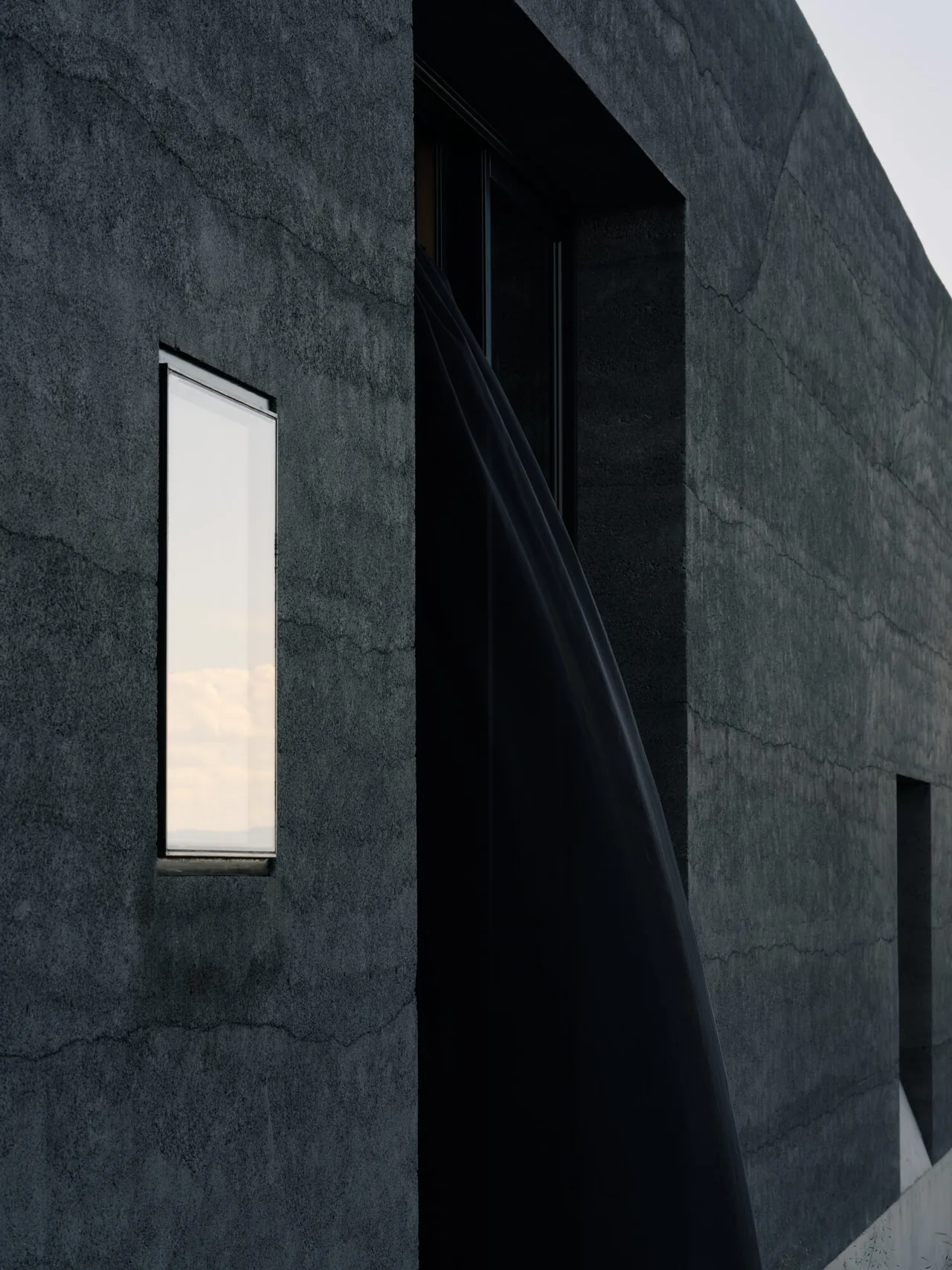 Dark concrete facade features a narrow vertical window and a recessed sliding glass door revealing interior shadow.