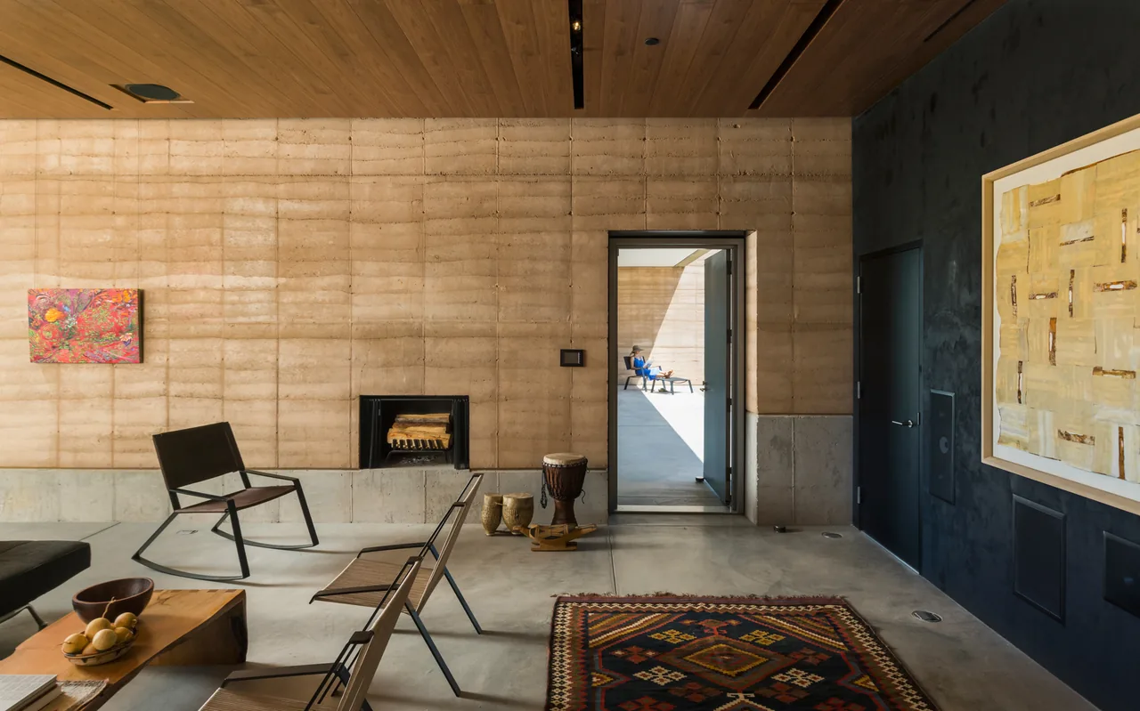 Living room in Tucson Mountain Retreat showcasing rammed earth walls and a view through an open door to an outdoor patio seating area.