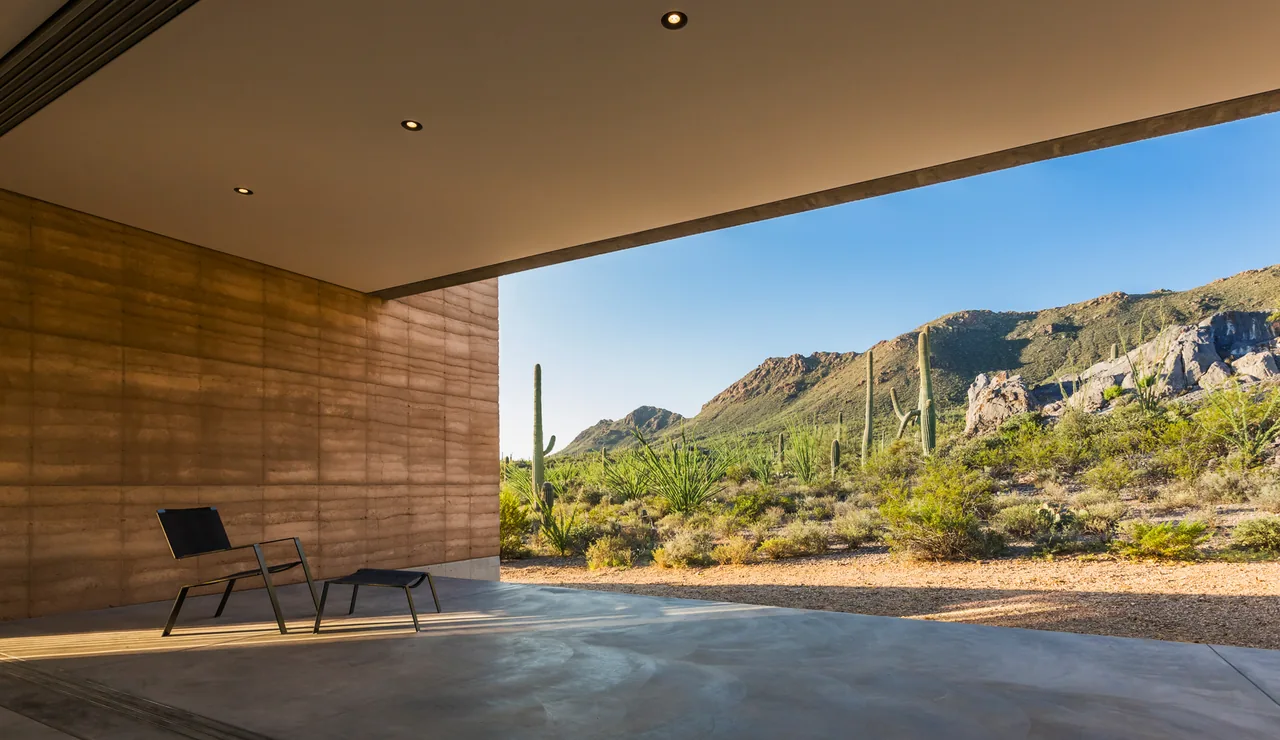 The Tucson Mountain Retreat features a covered patio with rammed earth walls offering views of desert vegetation and mountains.