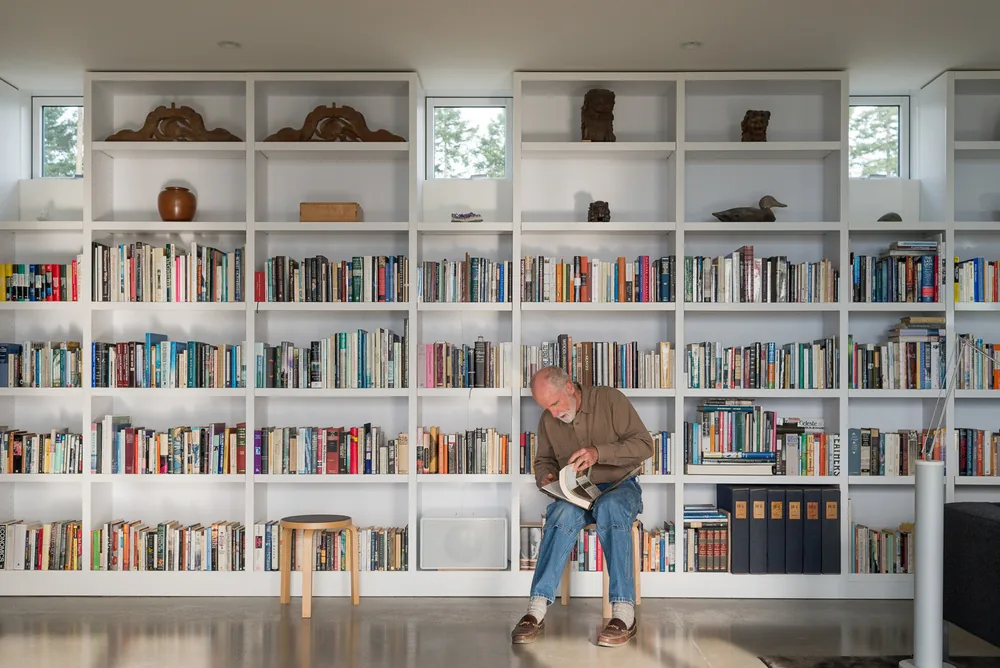 Interior view of a living or office space with a tall white built-in bookshelf and a man reading a book.