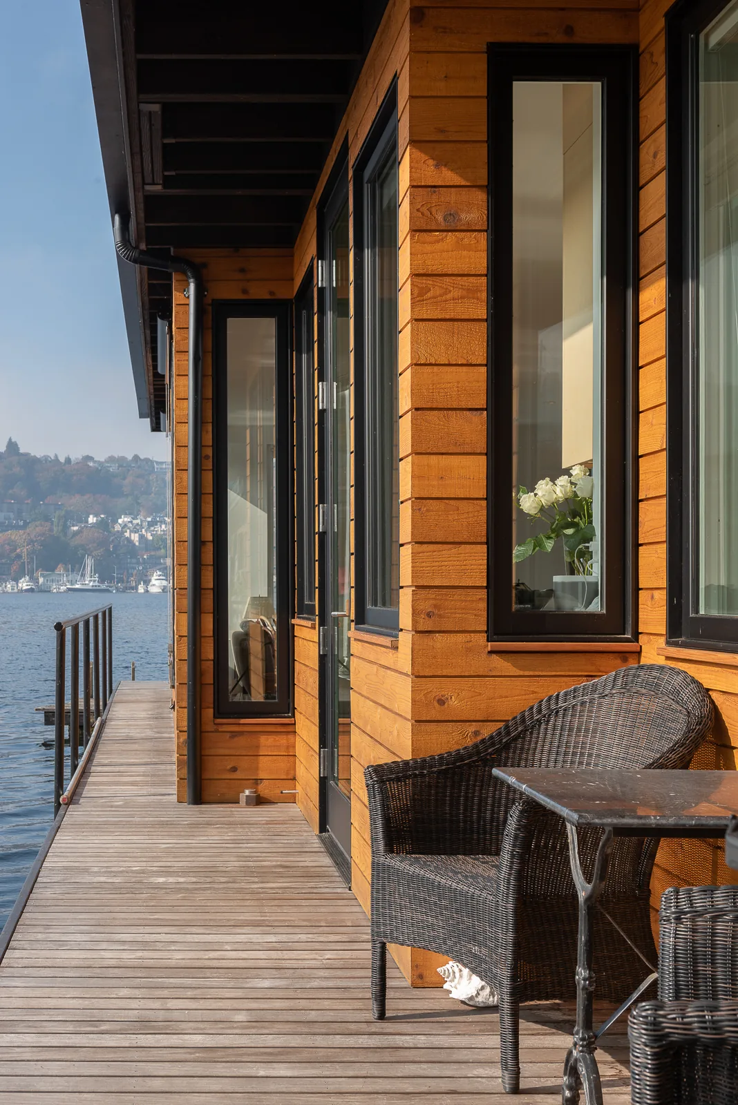 Exterior lakeside porch of a floating home with natural wood siding, black window frames, and outdoor seating.